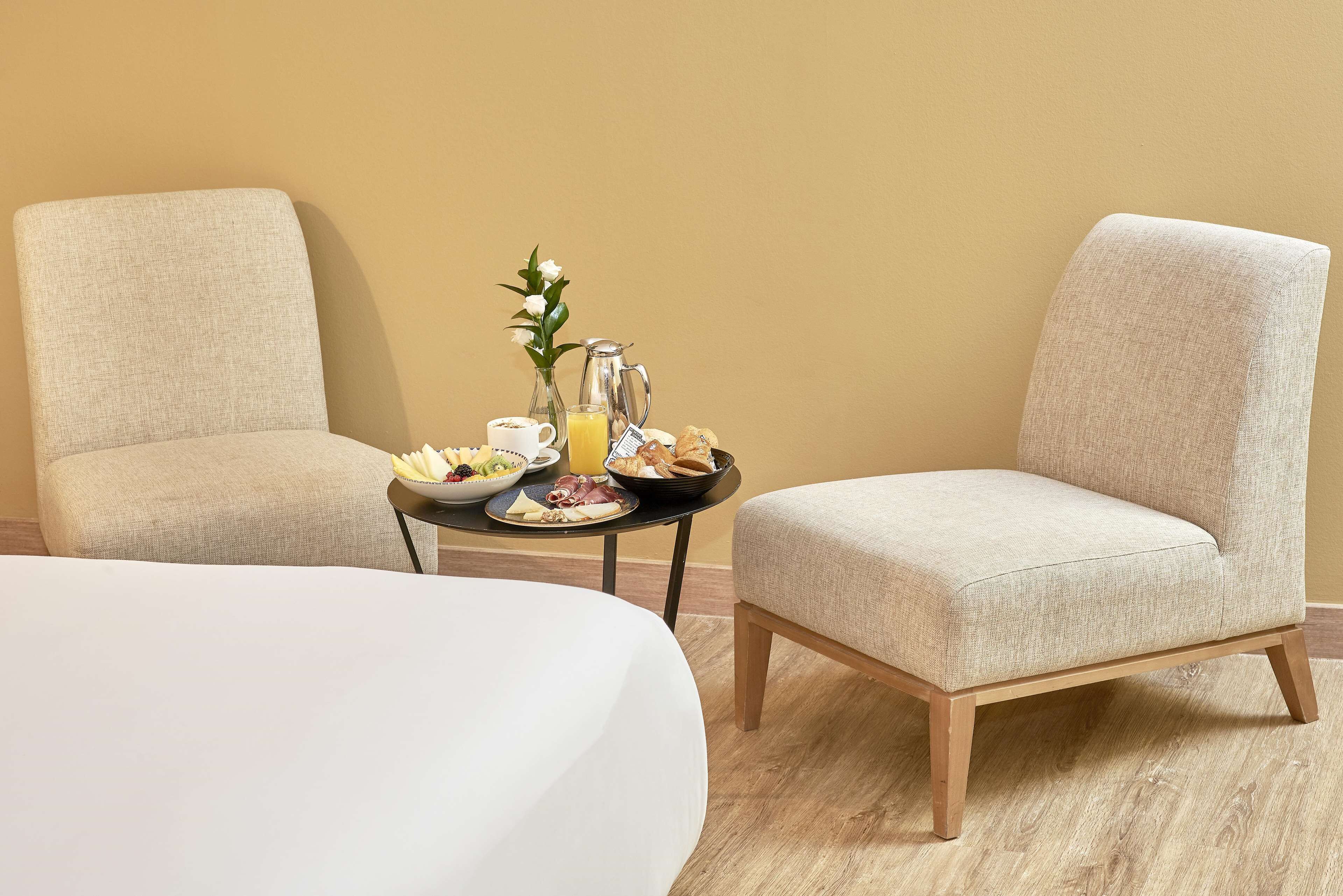 A hotel room with two beige armchairs, a small round table with breakfast items, and a white bed.