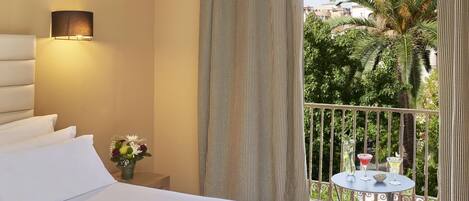 A hotel room with a bed, a nightstand with a vase of flowers, a balcony with a table and drinks, and a view of palm trees and buildings.