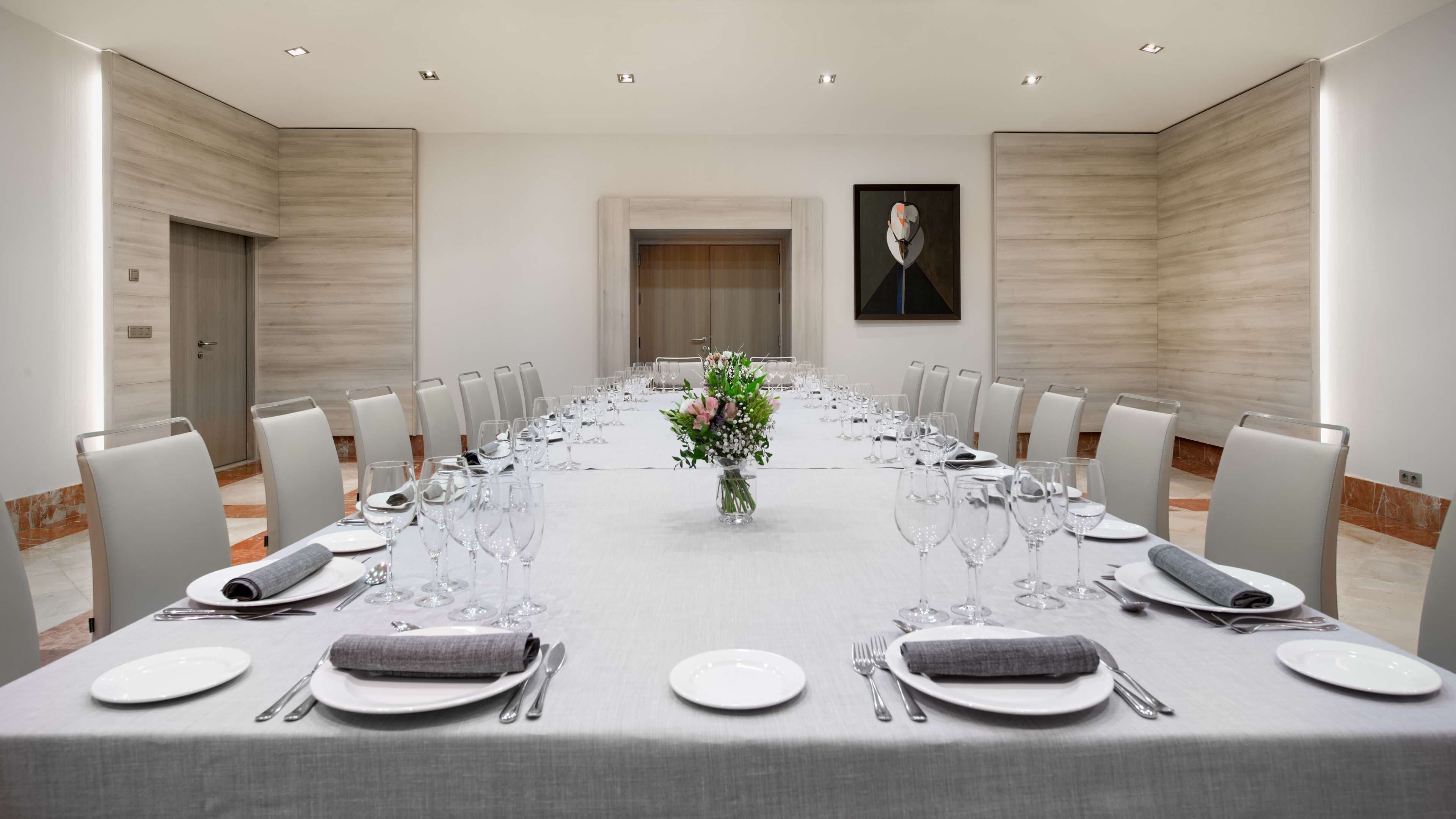 Ein Konferenzraum mit einem langen Tisch, der für ein Meeting gedeckt ist, umgeben von Stühlen, mit einer Blumenvase in der Mitte.