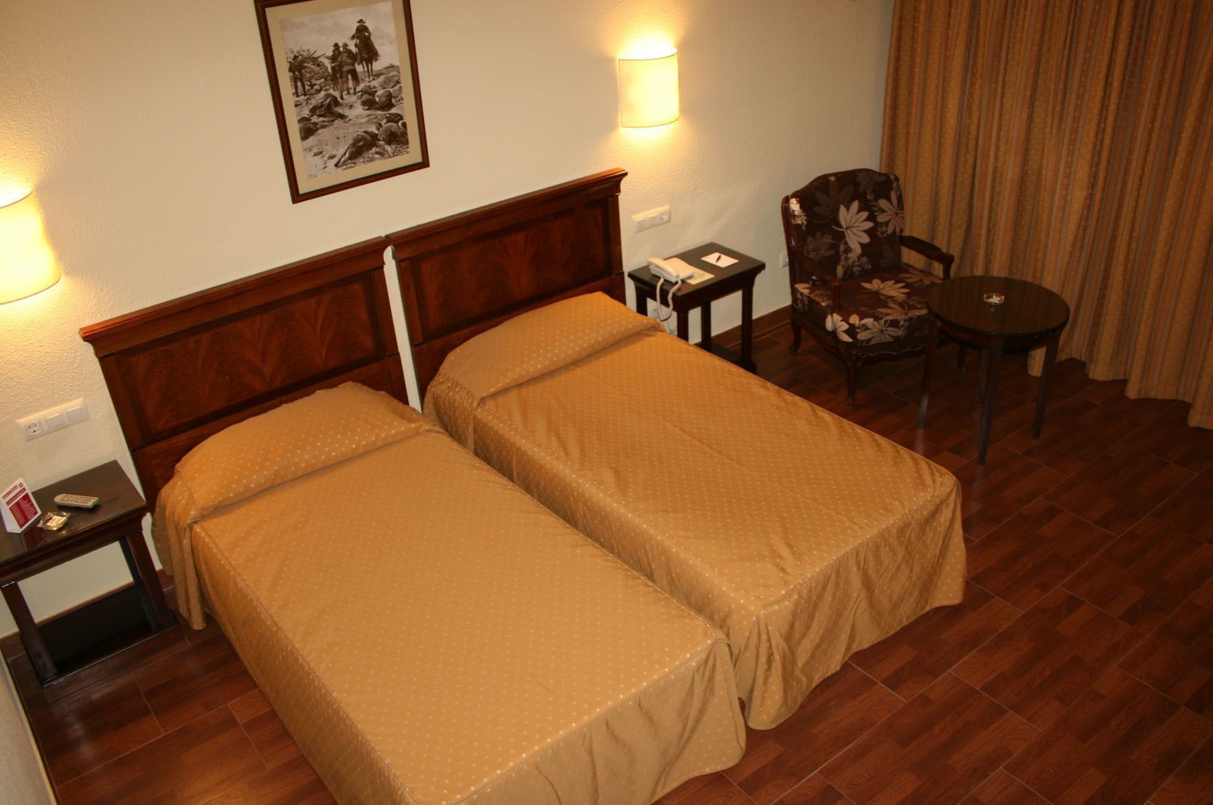 A hotel room with two single beds, a wooden headboard, a small table with a chair, a lamp, and a framed picture on the wall.