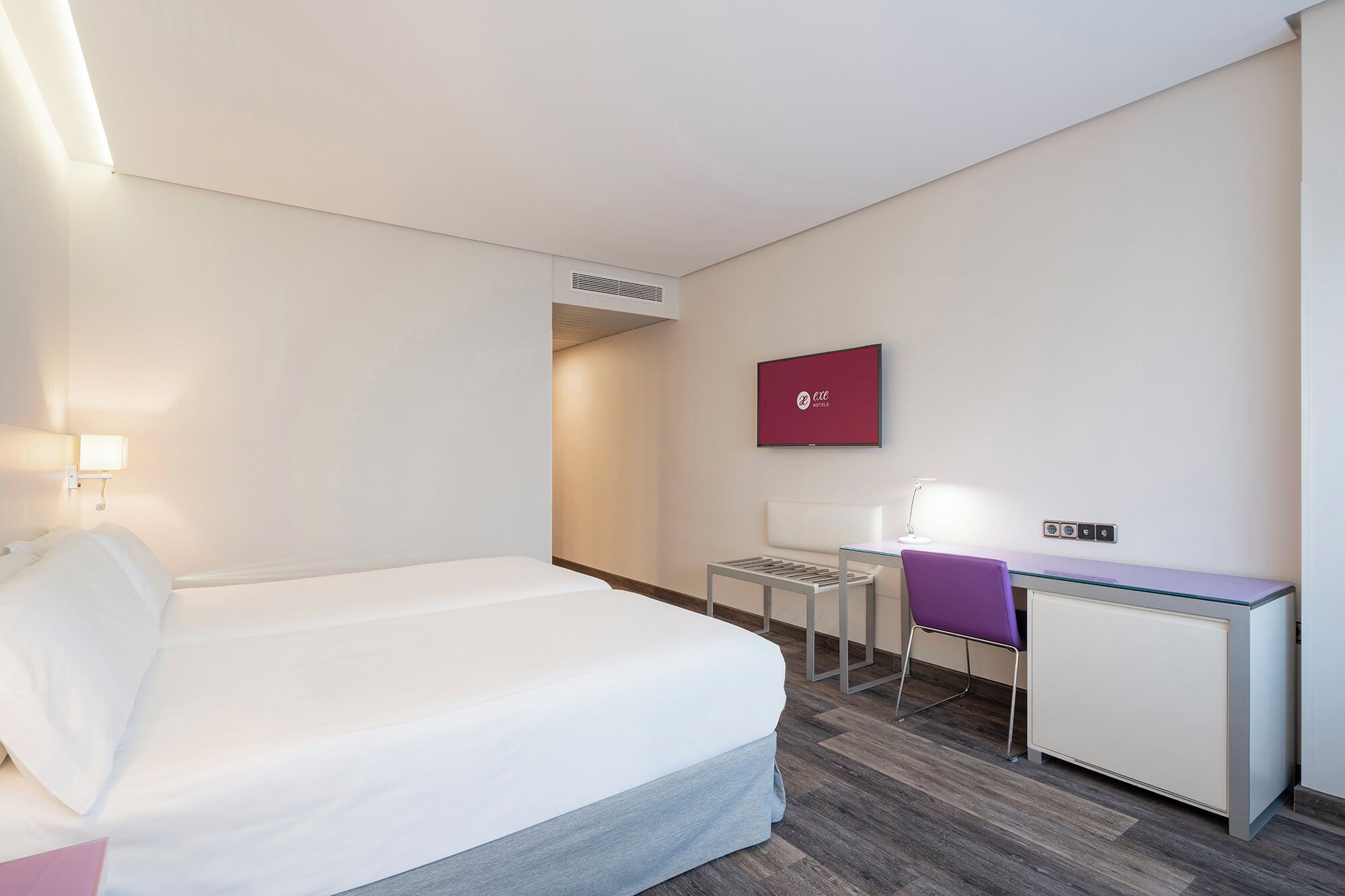 A hotel room with a bed, a desk, a chair, and a TV on the wall.