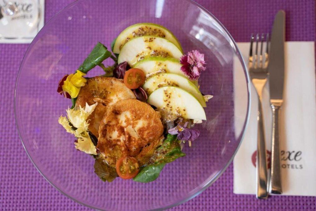 A clear glass bowl containing a salad with sliced apples, lettuce, and a piece of bread, placed on a purple tablecloth.