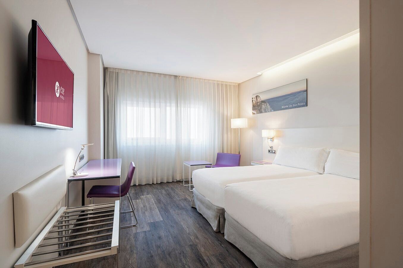 Habitación de hotel con cama, escritorio con silla, televisor montado en la pared y ventana con cortinas.