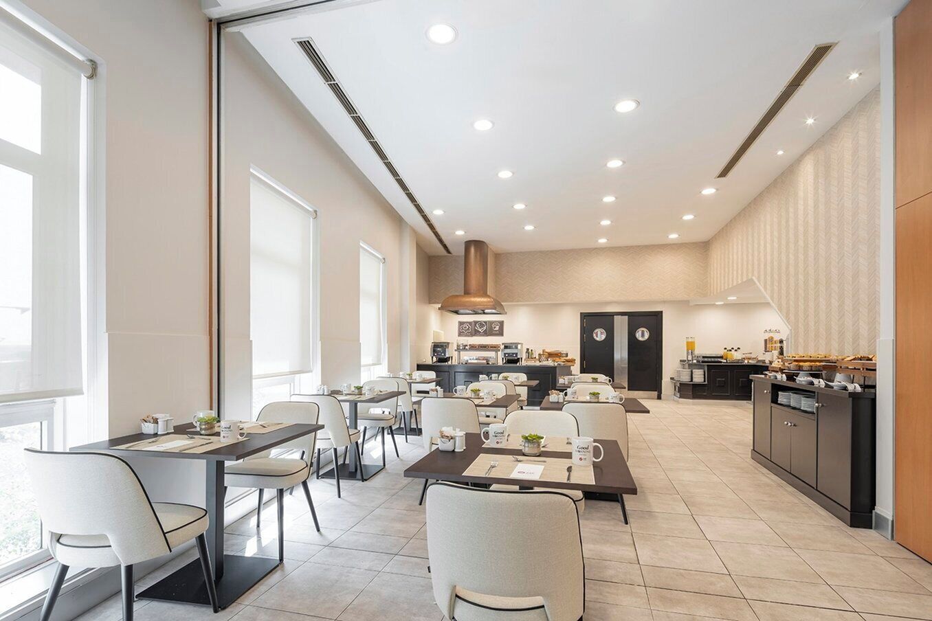 A modern dining area with multiple tables and chairs, a kitchen in the background, and a large window providing natural light.