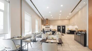 A modern dining area with multiple tables and chairs, a kitchen in the background, and a large window providing natural light.