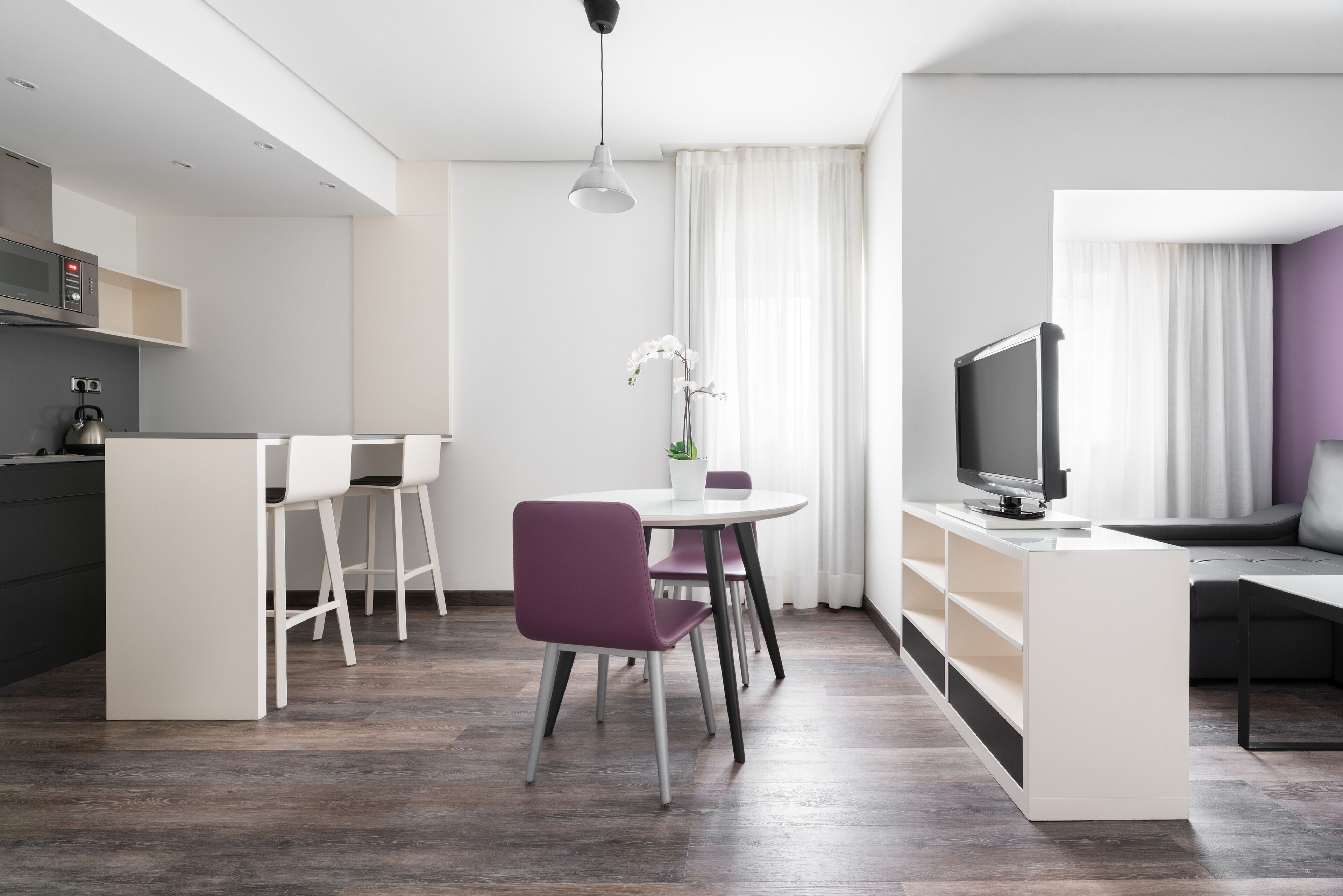 Una cocina moderna con microondas, tetera y una pequeña isla con asientos. Hay un comedor con una mesa redonda y sillas moradas. Un televisor está montado en la pared y hay una orquídea en una maceta sobre la mesa.