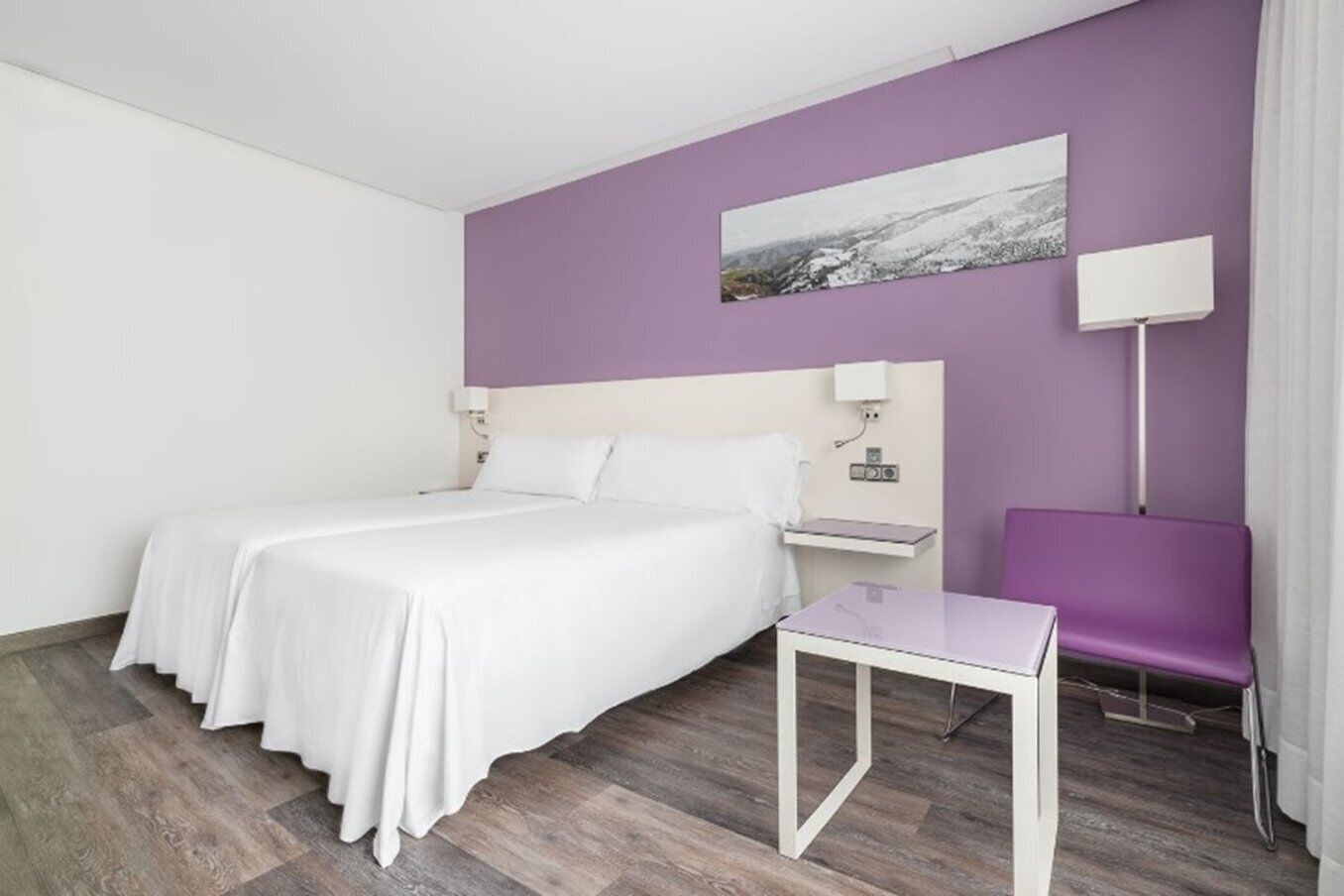 Et hotelværelse med en seng, et lille bord, en stol, et natbord og en lampe fastgjort til væggen.