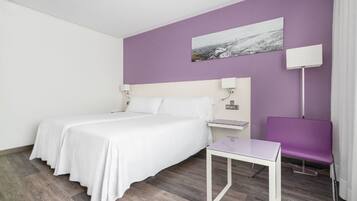 Et hotelværelse med en seng, et lille bord, en stol, et natbord og en lampe fastgjort til væggen.