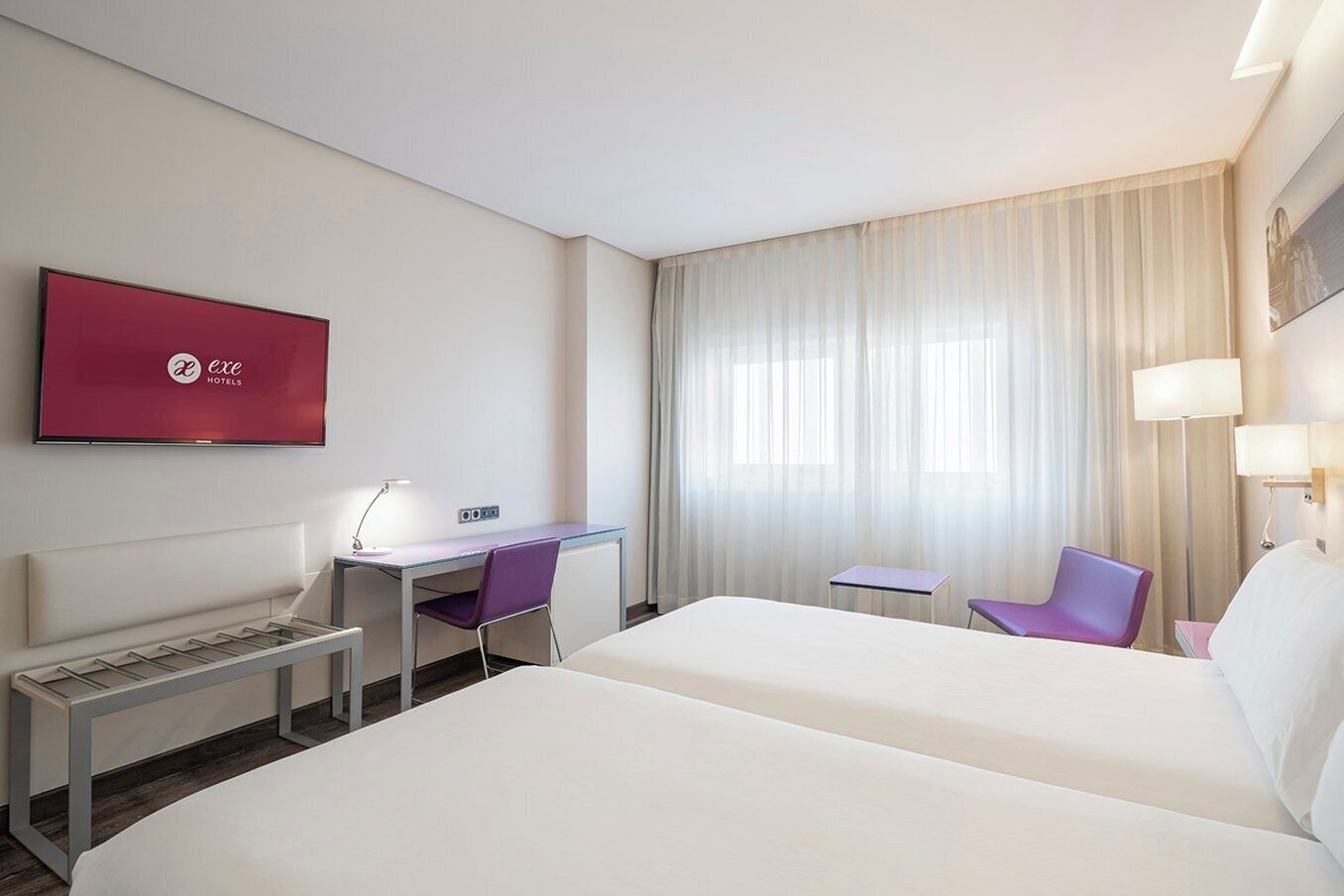 A hotel room with two beds, a desk, a chair, a TV, and a window with curtains.