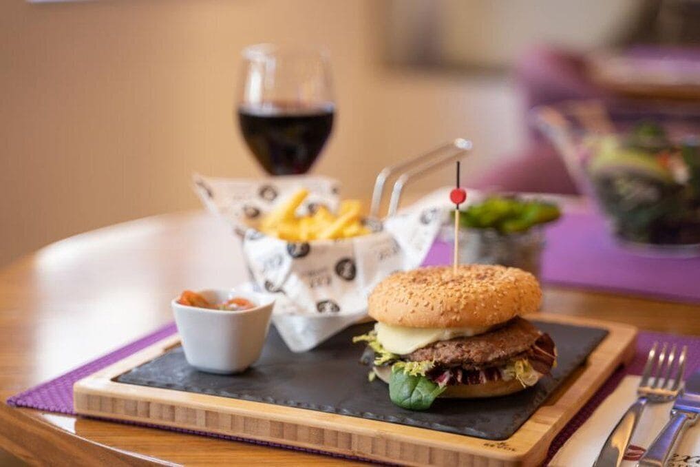 Una hamburguesa con papas fritas y un tazón pequeño de salsa sobre una tabla de madera, con una copa de vino tinto al fondo.