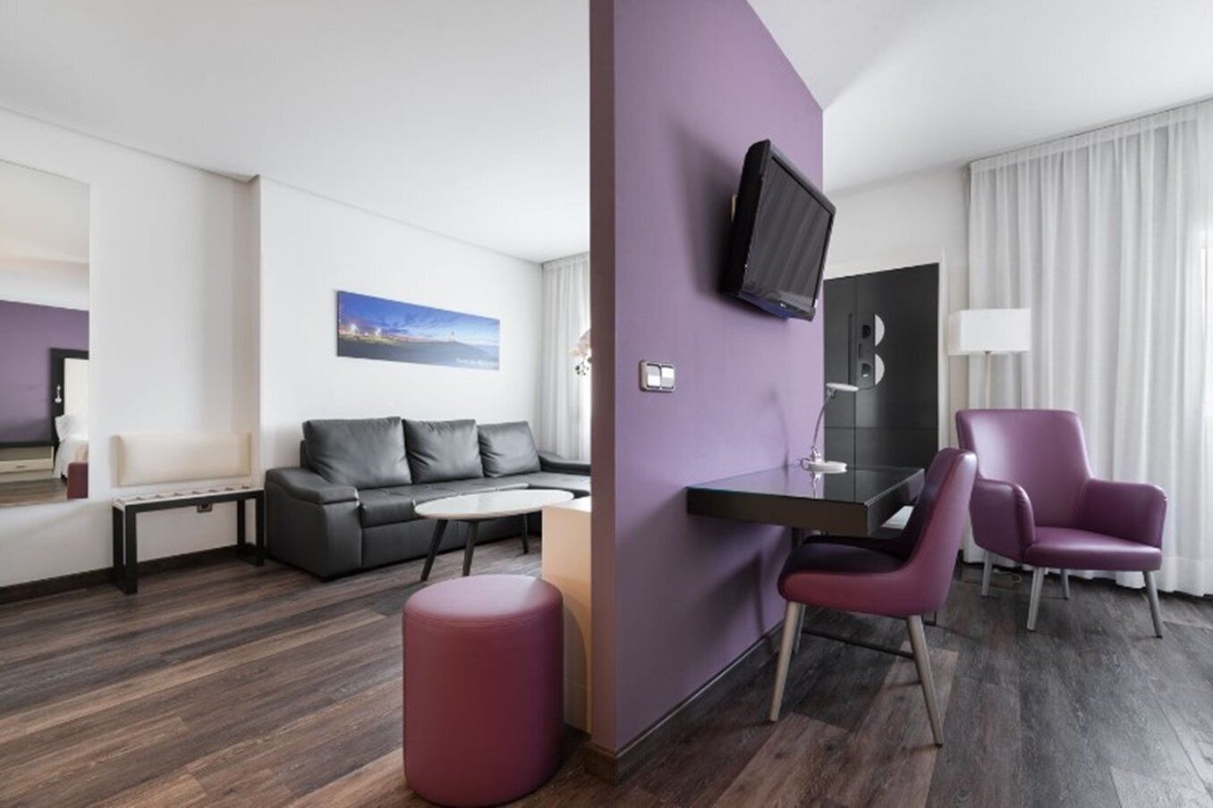 Una habitación de hotel moderna con un sofá gris, una mesita redonda pequeña, un sillón morado, un televisor montado en la pared y una ventana con cortinas blancas.