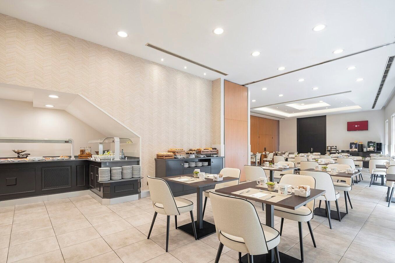 A modern hotel dining area with multiple tables and chairs, a buffet setup, and a staircase leading to an upper level.