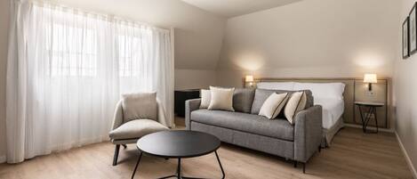 A modern hotel room with a grey sofa, a round black table, a single armchair, and a bed with a headboard. There are two bedside lamps and a small table with a lamp.