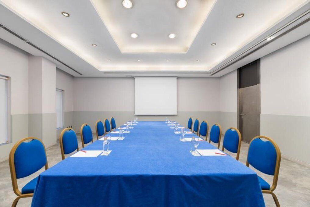 Une salle de conférence avec une longue table, des chaises bleues et un écran de projection.
