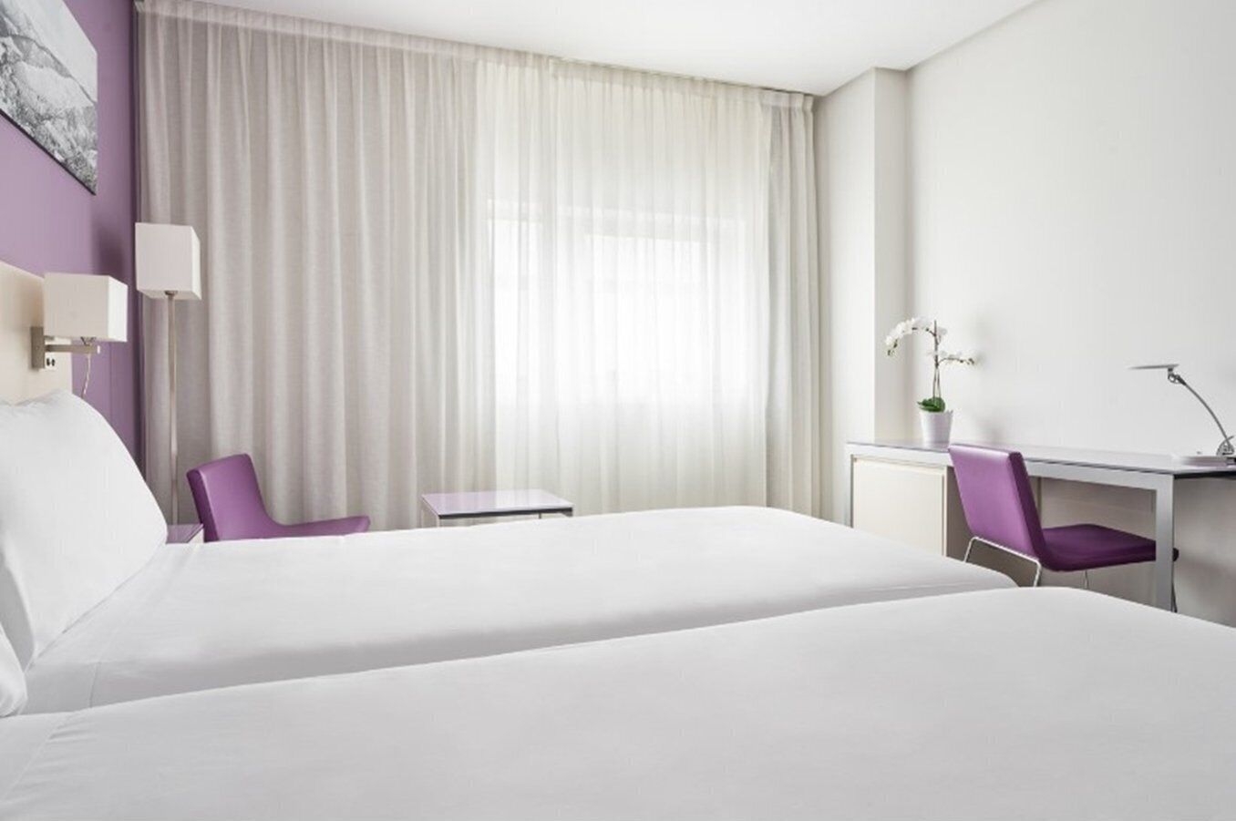 A hotel room with a large bed, a desk with a chair, a small table, and a window with sheer curtains.