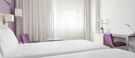 A hotel room with a large bed, a desk with a chair, a small table, and a window with sheer curtains.