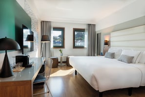 Una habitación de hotel moderna con una cama grande, un escritorio con lámpara, una silla, una mesita con una planta y dos ventanas con cortinas.