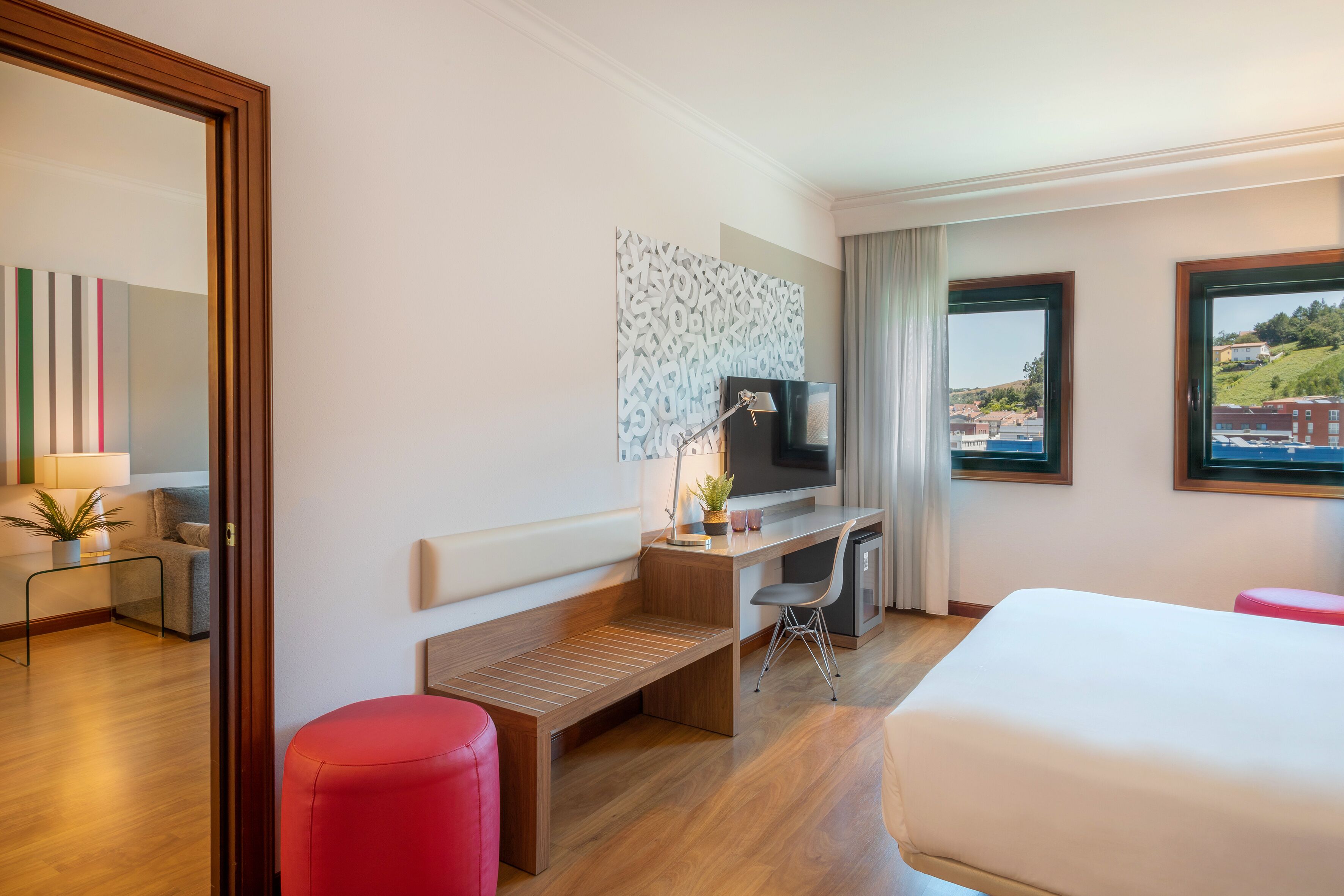 A hotel room with a bed, a desk with a chair, a small table with a lamp, a red ottoman, and a large window with a view of buildings and hills.
