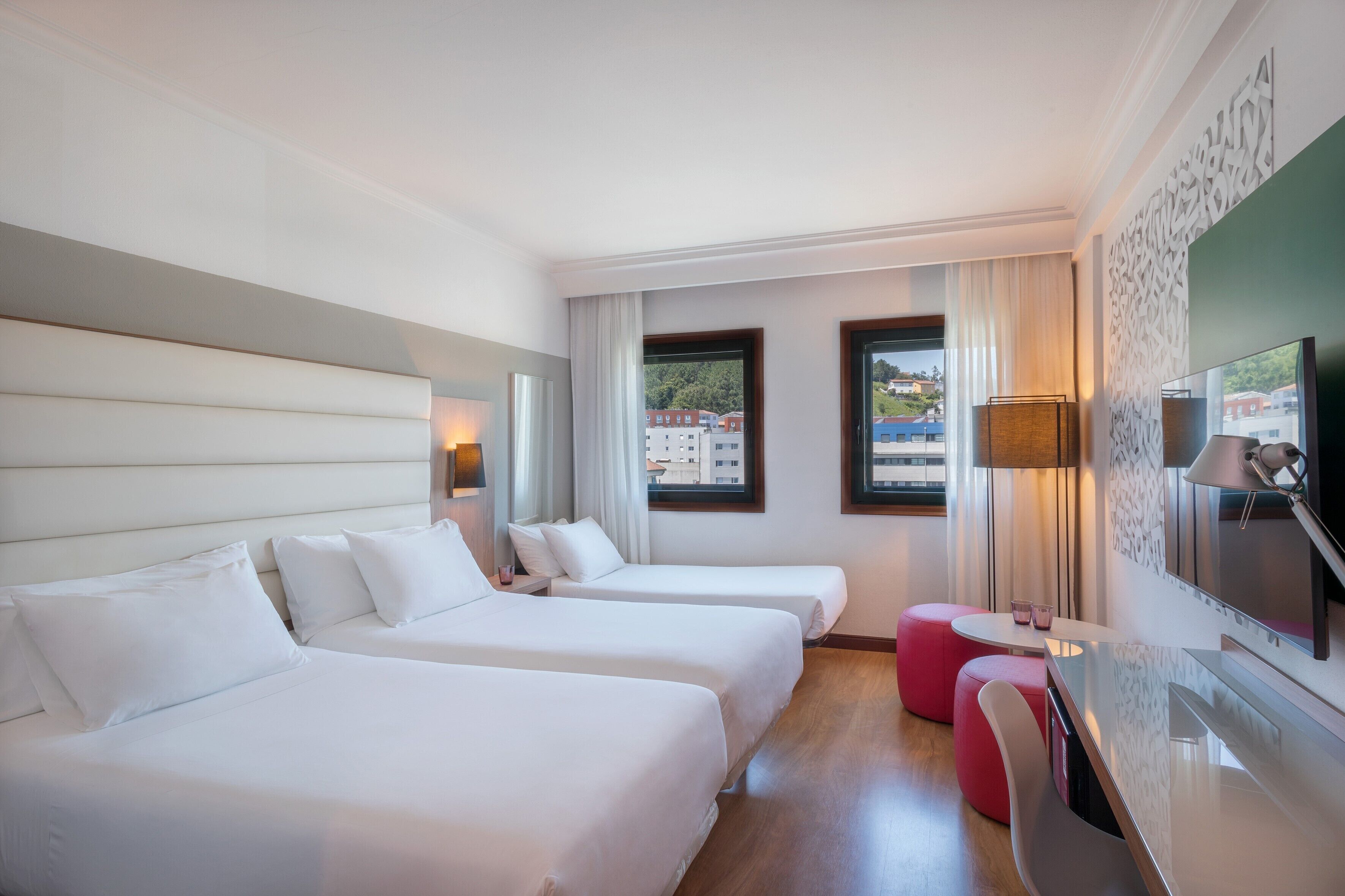 Habitación de hotel con dos camas, televisión, escritorio y vistas a edificios.