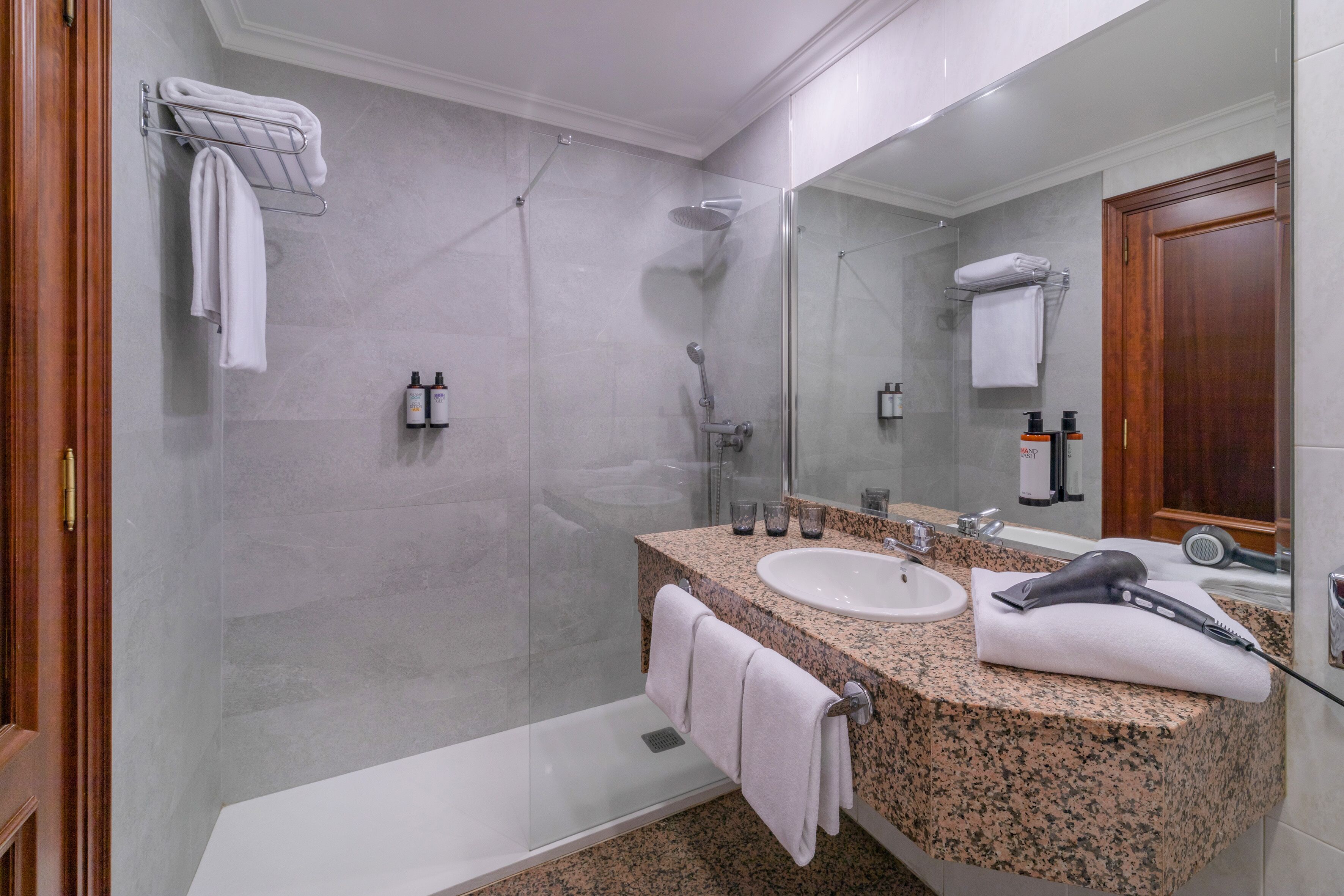 Une salle de bain moderne dotée d’un comptoir en granit, d’un lavabo blanc, d’un sèche-cheveux et d’une douche à parois de verre.