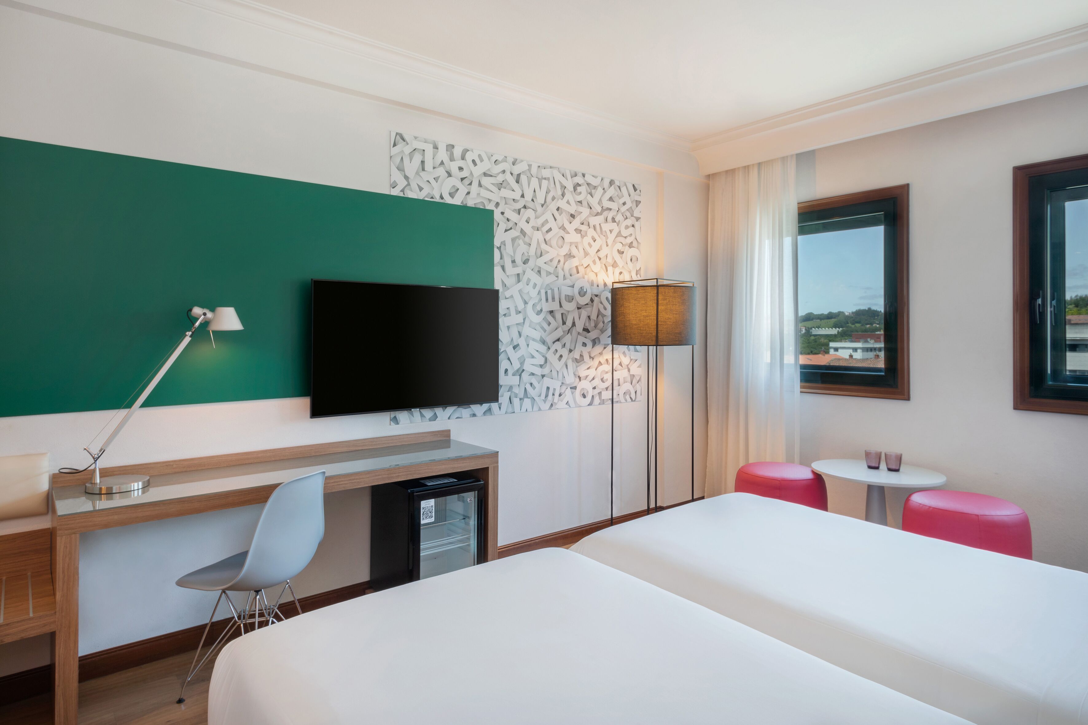 Habitación de hotel con cama, escritorio, silla, televisor, lámpara y vistas a la ciudad desde la ventana.