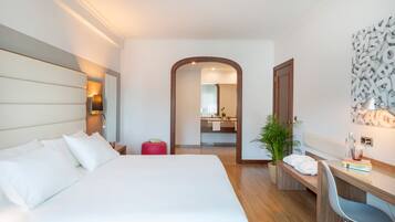 Una habitación de hotel con una cama grande, un escritorio y una silla. Hay un espejo, una planta y un taburete rojo.