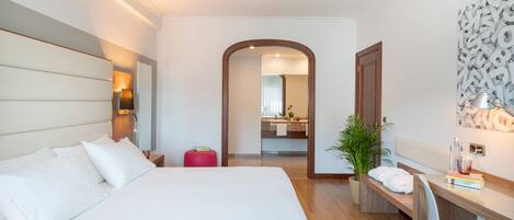A hotel room with a large bed, a desk, and a chair. There is a mirror, a plant, and a red stool.