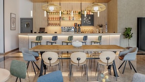 A modern bar area with a long wooden table, multiple chairs, and a variety of bottles on shelves.