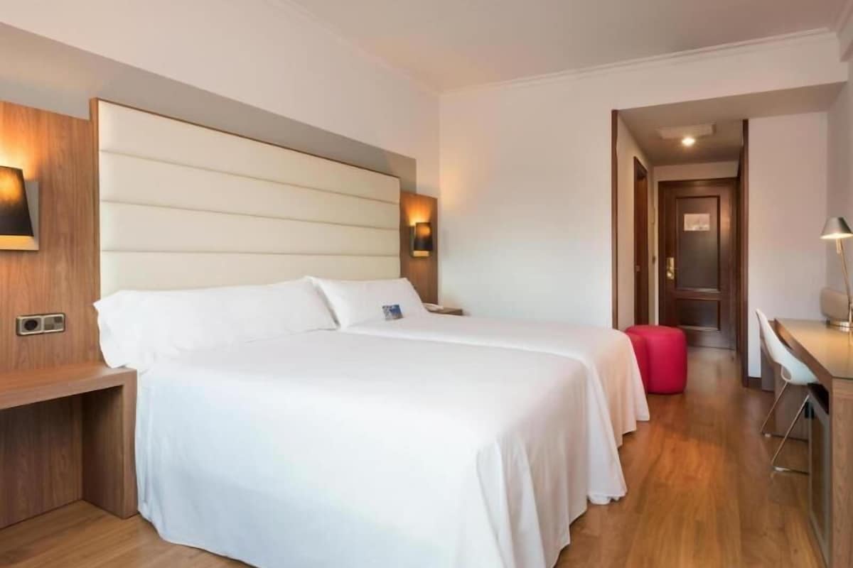 Une chambre d’hôtel avec deux lits, une tête de lit en bois, des lampes de chevet, un bureau et une chaise rouge.