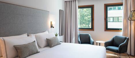 Ein Hotelzimmer mit einem großen Bett, zwei Sesseln, einem kleinen Tisch und einem Fenster mit Vorhängen.