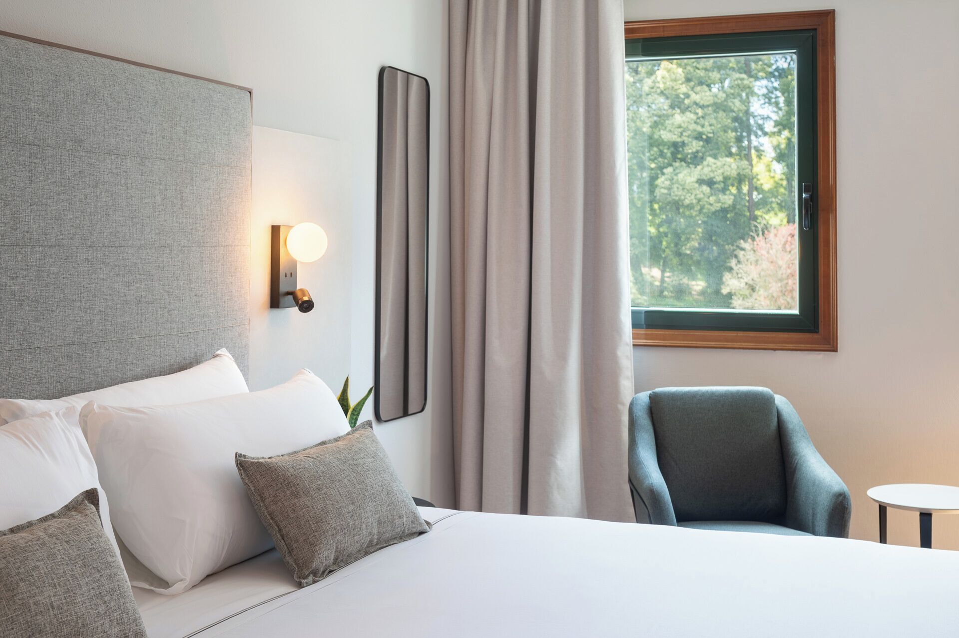 Habitación de hotel con cama, una silla, ventana con vista a árboles y lámpara de pared.