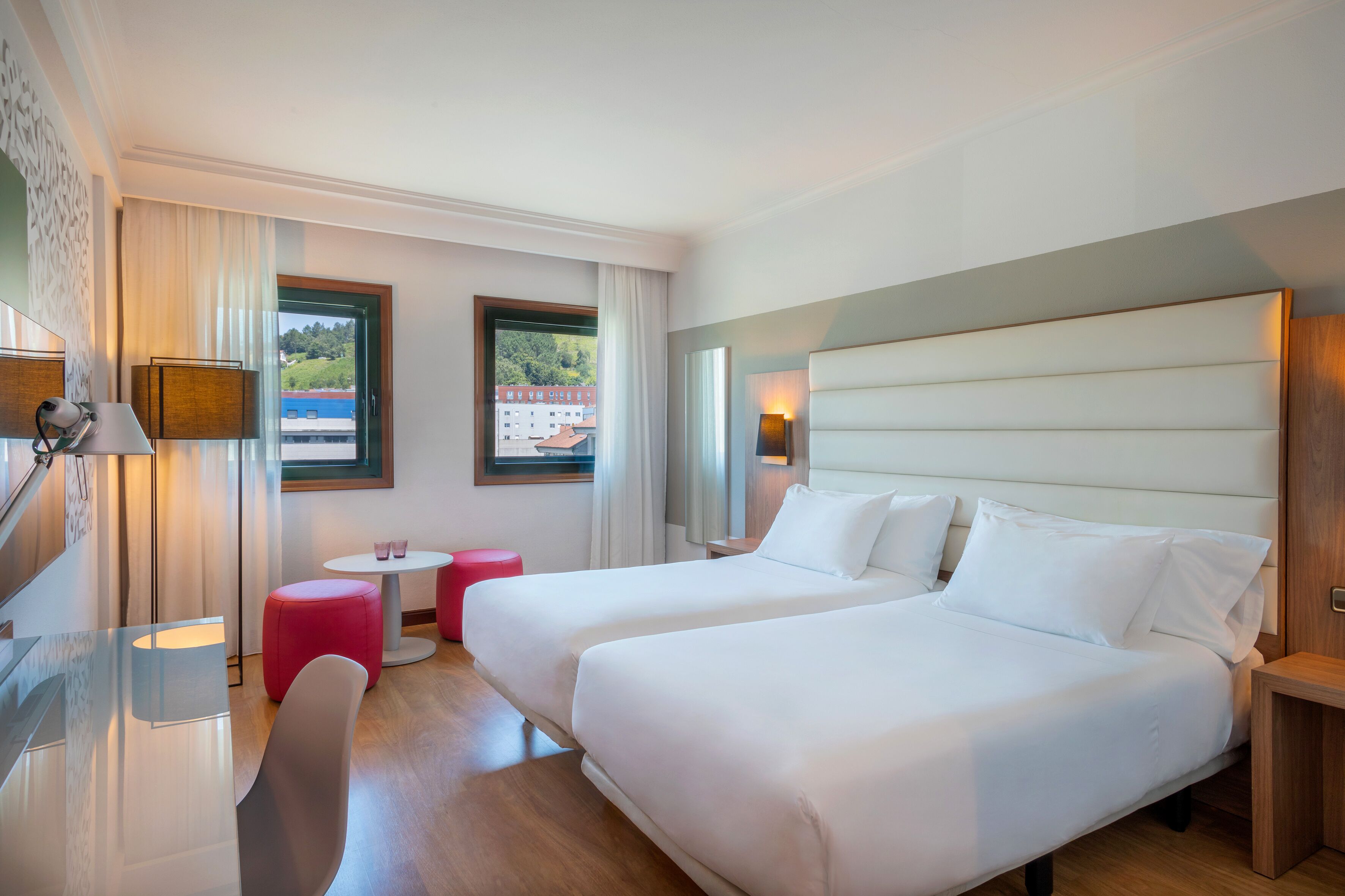 A hotel room with two beds, a desk, a chair, a small table, and a window with a view of buildings and greenery.