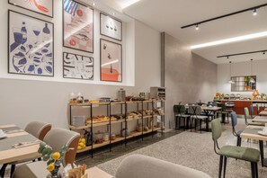 Ein moderner Essbereich mit einer langen Theke, mehreren Tischen und Stühlen. Regale mit Lebensmitteln und eine Wand mit gerahmten Kunstwerken sind ebenfalls vorhanden.
