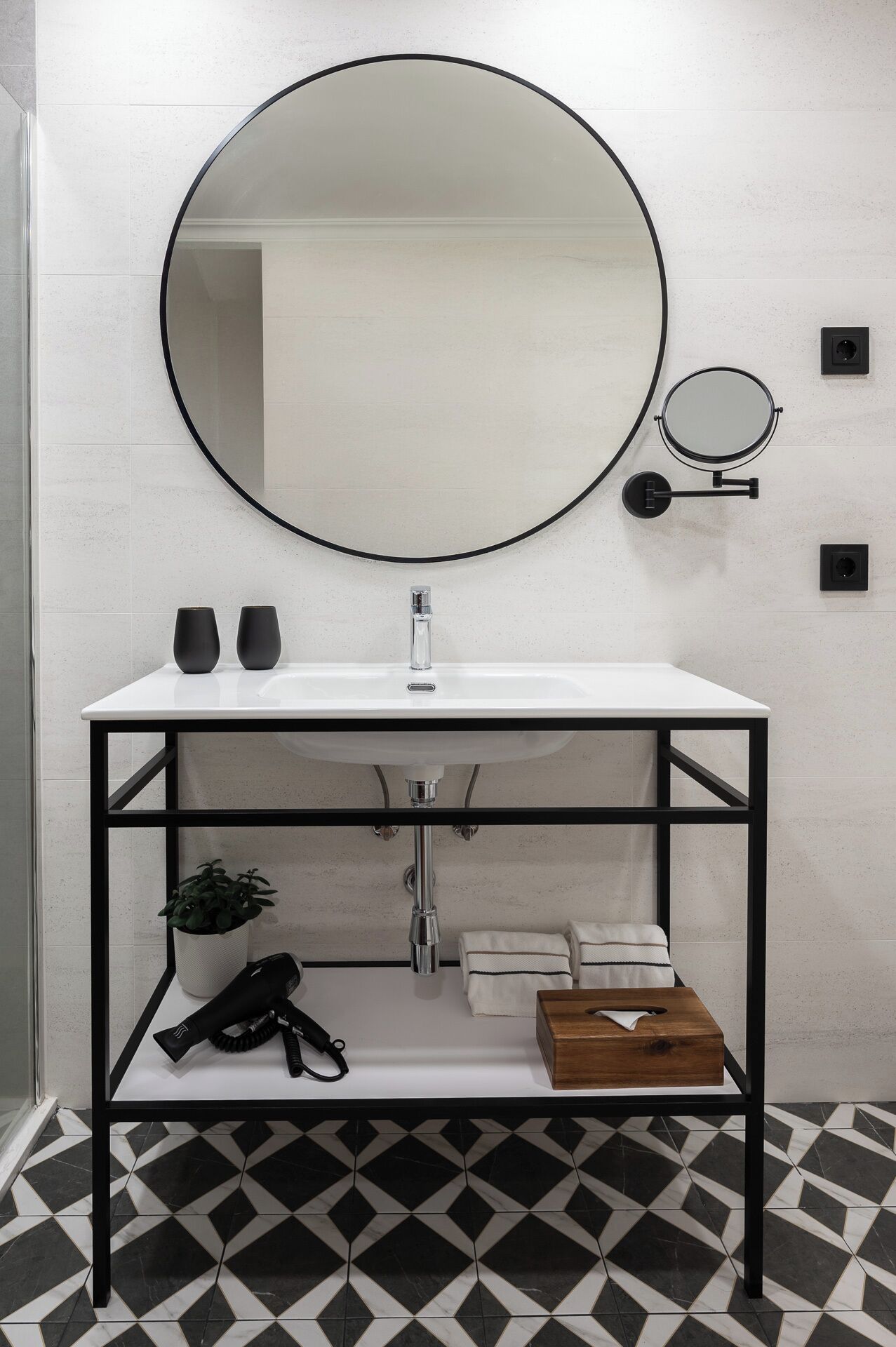 Een badkamer met een witte wasbak, een zwart metalen frame, een ronde spiegel, een kleine ronde spiegel en een potplant.