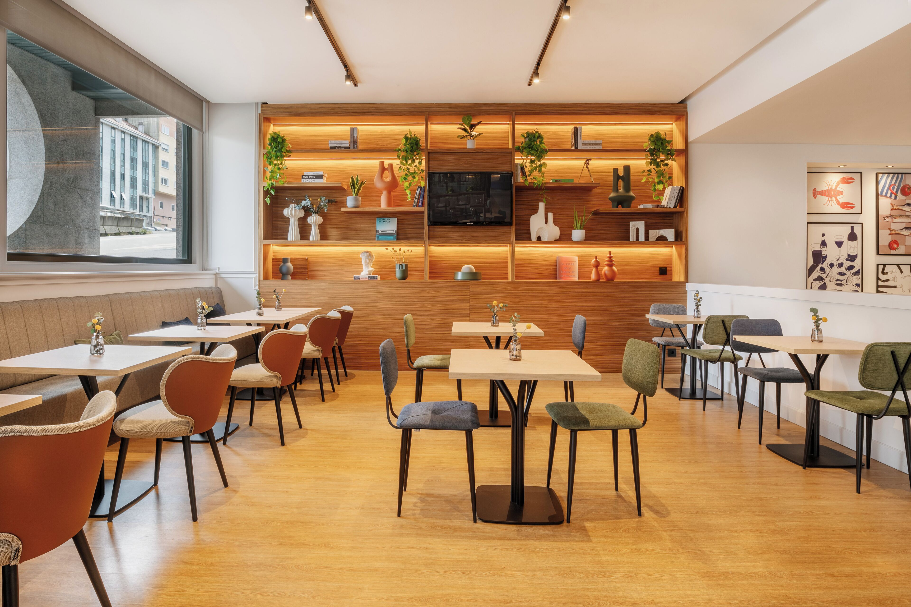 Ein modernes Restaurant mit Holztischen und -stühlen, einem an der Wand befestigten Fernseher sowie Regalen mit Pflanzen und Büchern.