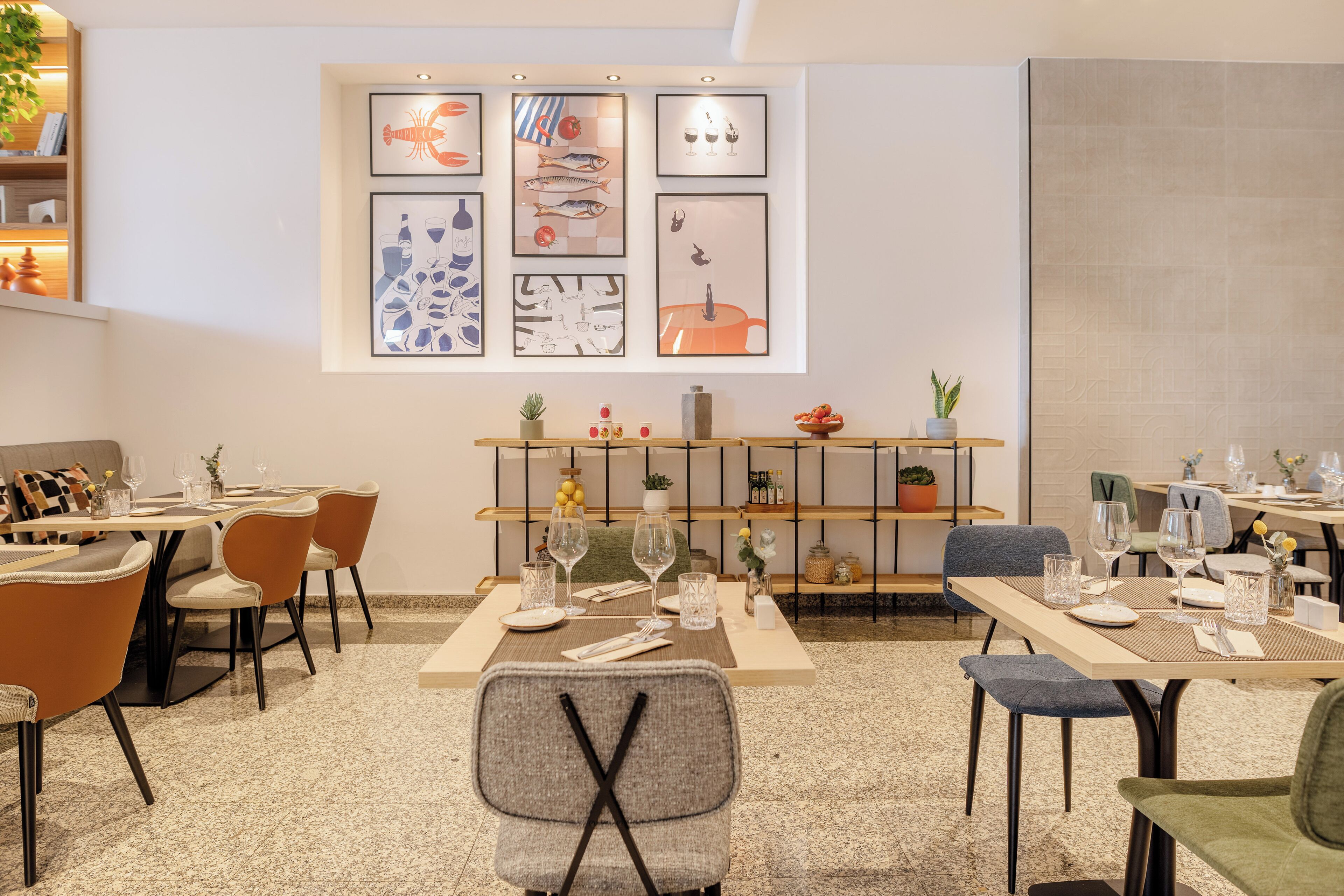 A dining area with multiple tables and chairs, framed artwork on the wall, and a plant on the shelf.