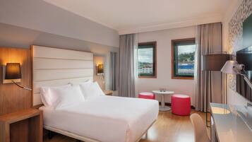 Quarto de hotel moderno com uma cama grande, mesas de cabeceira, uma escrivaninha, duas cadeiras e uma pequena mesa com um banquinho vermelho.