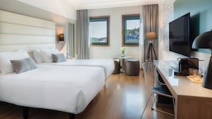 A hotel room with two beds, a desk, a TV, and a view of the city.