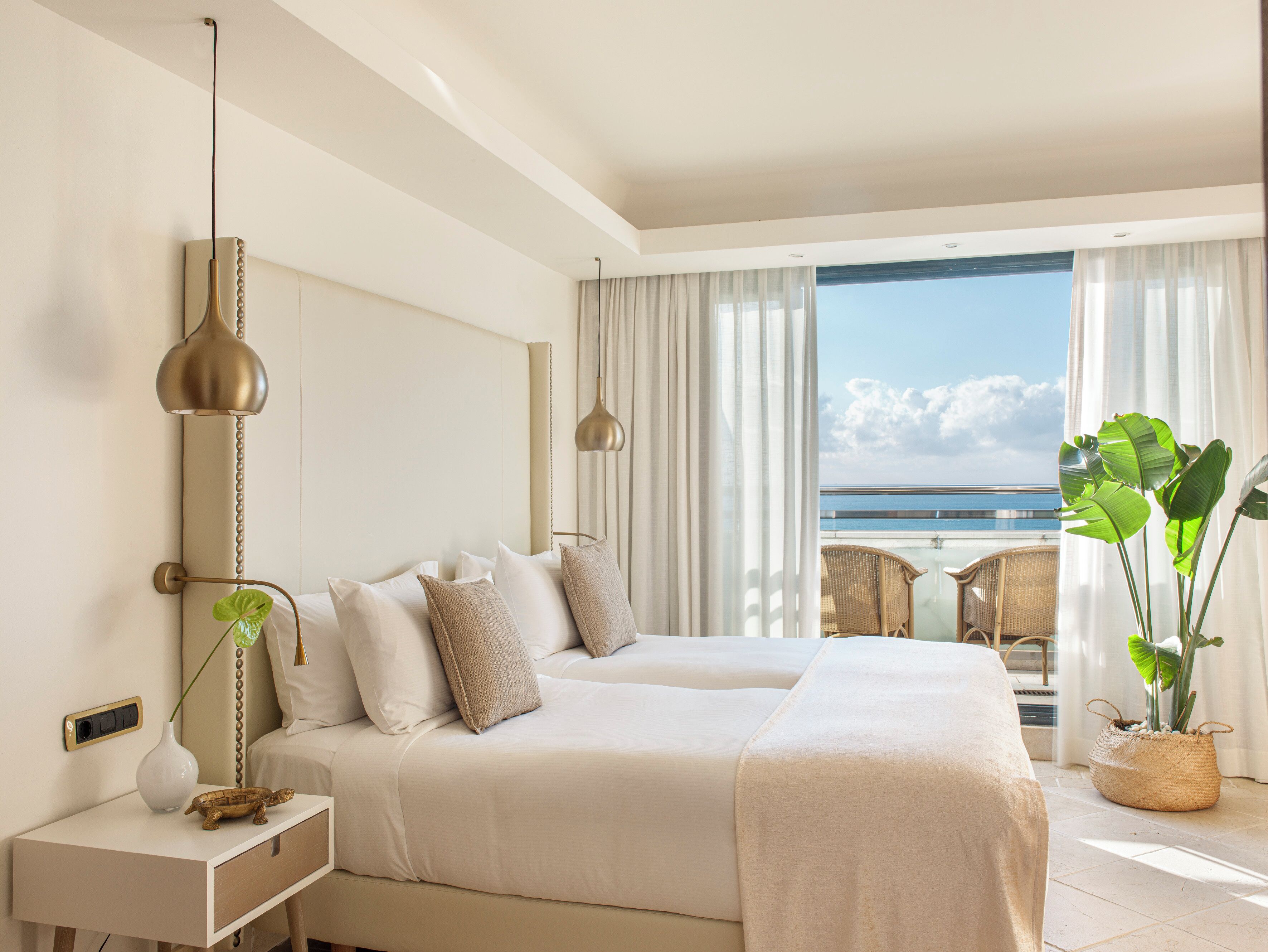 Habitación de hotel con una cama grande, una mesita de noche con lámpara, vista al océano y un balcón con una silla.