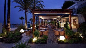 Un patio al aire libre con mobiliario de mimbre y suelo de piedra, rodeado de palmeras y plantas, bajo una pérgola de madera.
