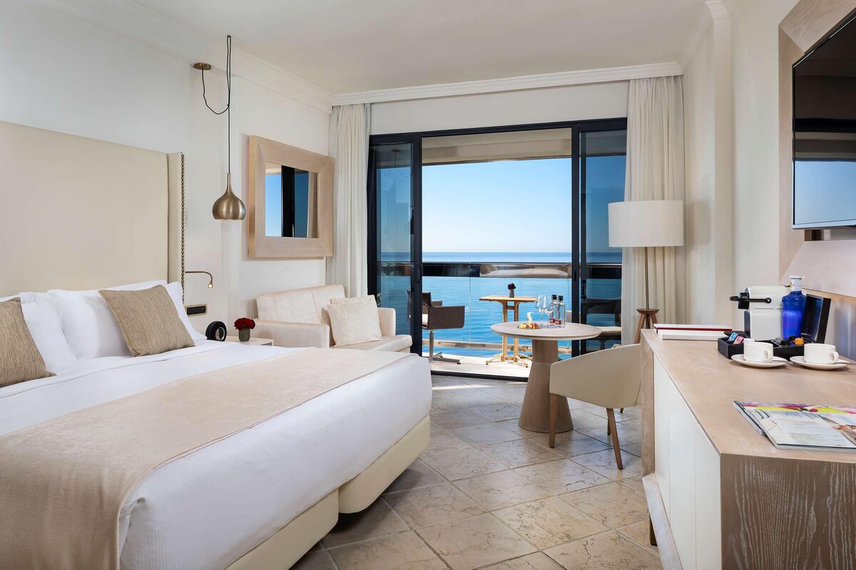 Une chambre d’hôtel avec un grand lit, un bureau, une chaise, une table et une vue sur la mer, visible à travers une porte vitrée.