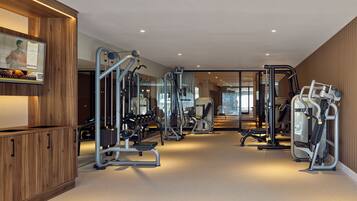 Un gimnasio moderno con diversas máquinas de ejercicio, un mostrador de recepción de madera y un espejo grande que refleja el espacio.