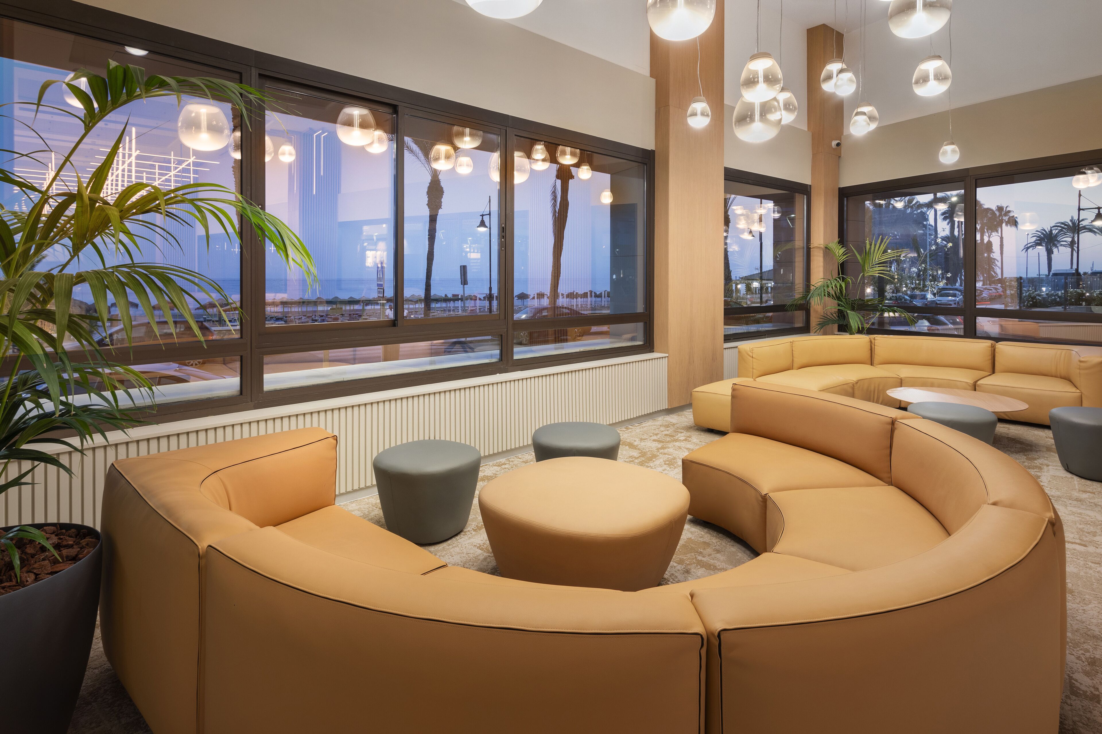 A modern lounge area with a curved sofa, round ottomans, and large windows offering city views.