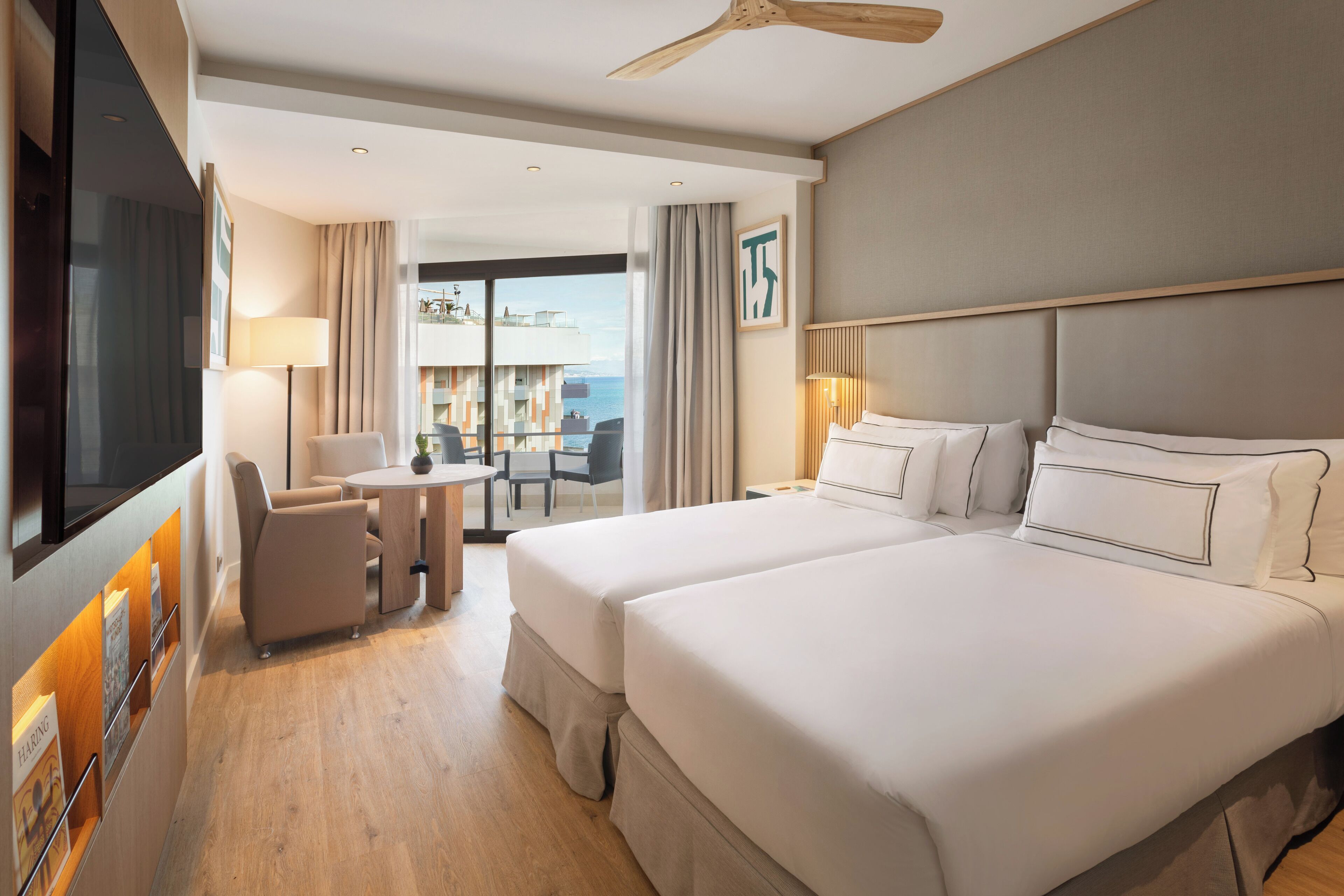 Ein Hotelzimmer mit zwei Betten, einem Fernseher, einem Schreibtisch mit Stuhl und einem Balkon mit Aussicht.