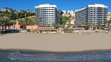 Een hotel aan het strand met meerdere hoge gebouwen, een zandstrand en palmbomen.