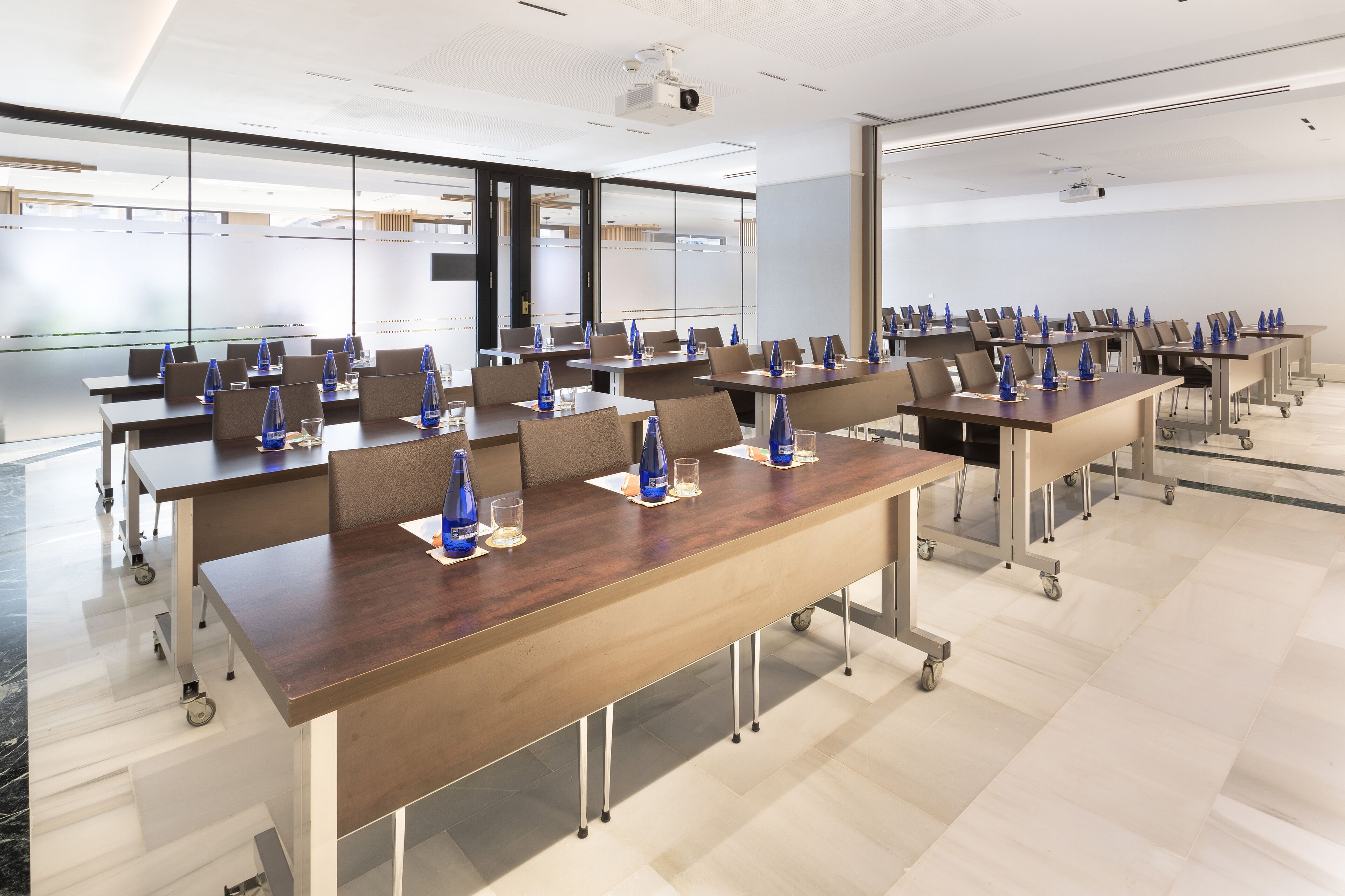 Un salón de conferencias con varias mesas, cada una con sillas y botellas de agua azules.
