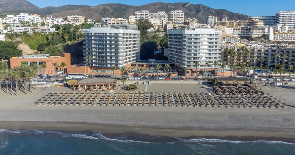 Un hotel frente al mar con varias edificaciones, una playa con tumbonas y montañas al fondo.