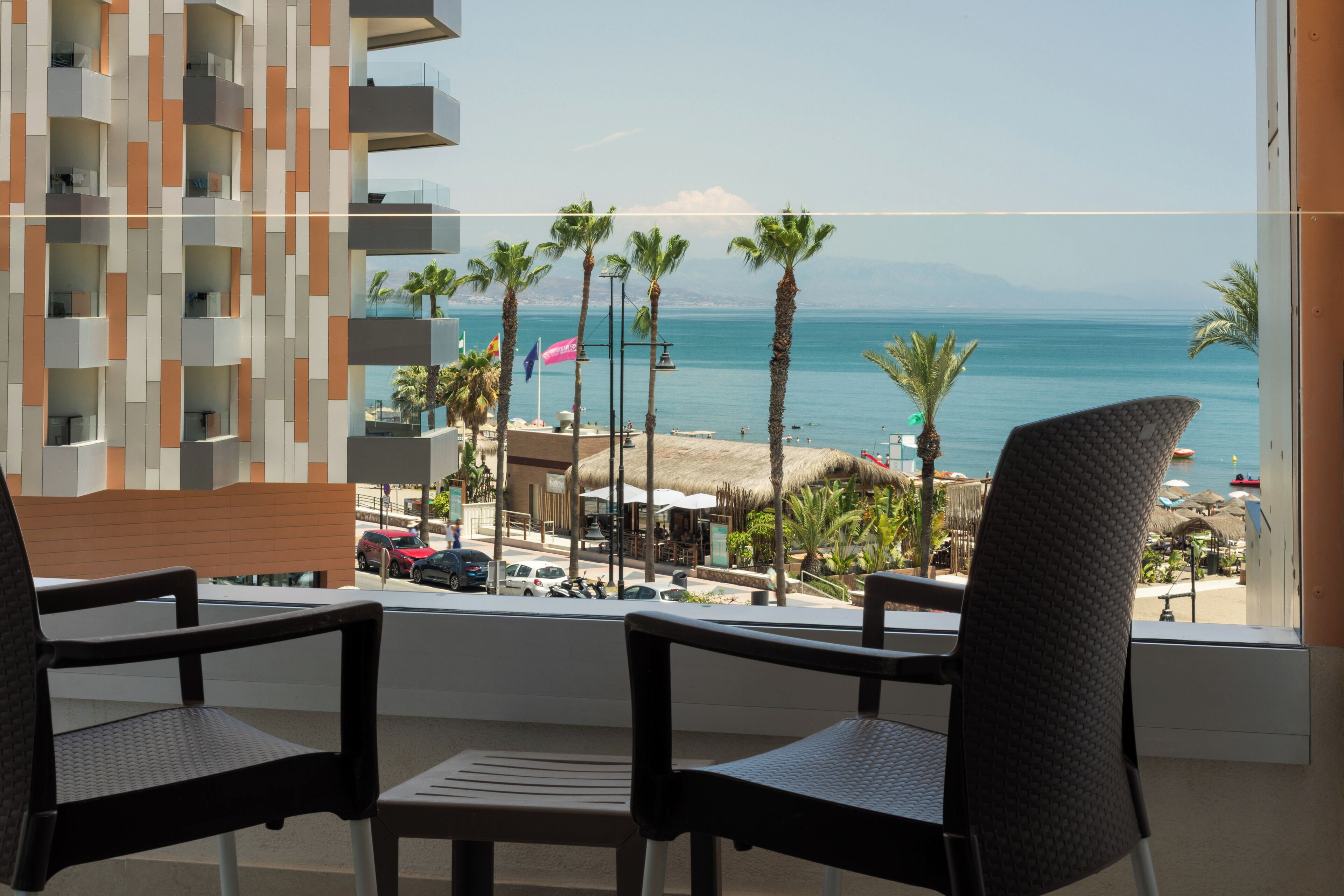 Ein Balkon mit zwei Stühlen, der Blick auf einen Strand mit Palmen und das Meer freigibt.