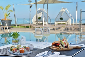 A dining table with sushi, two glasses of white wine, and a sandwich by a poolside.