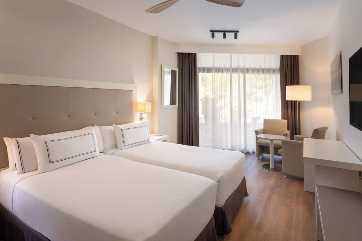 Ett hotellrum med två sängar, ett skrivbord, en stol, en TV och ett fönster med gardiner.